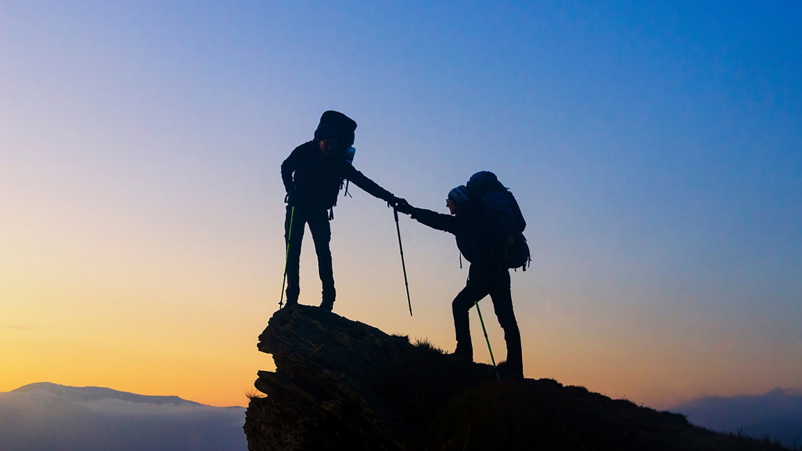 Two people at the summit of a mountain, one giving the other one a hand to the top