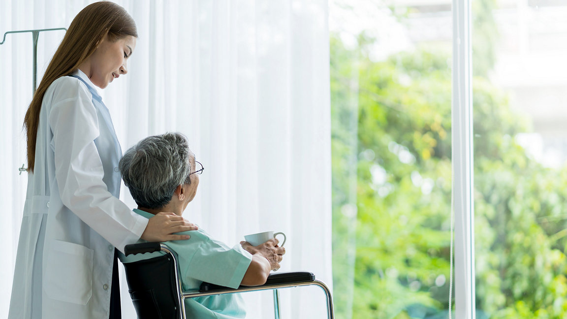 Female hospital staff with elderly patient in wheelchair looking out of floor-to-ceiling window with view of greenery