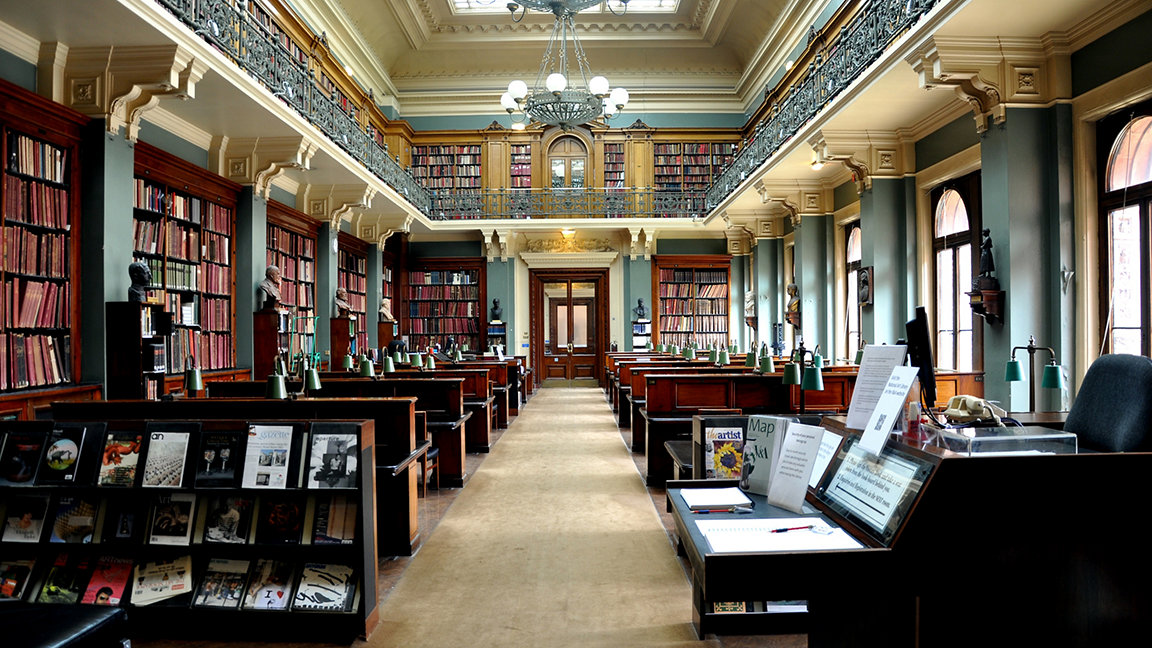 An interior of a Victorian library, with shelves of books and magazines, with further shelving on a gallery with ornate iron railings