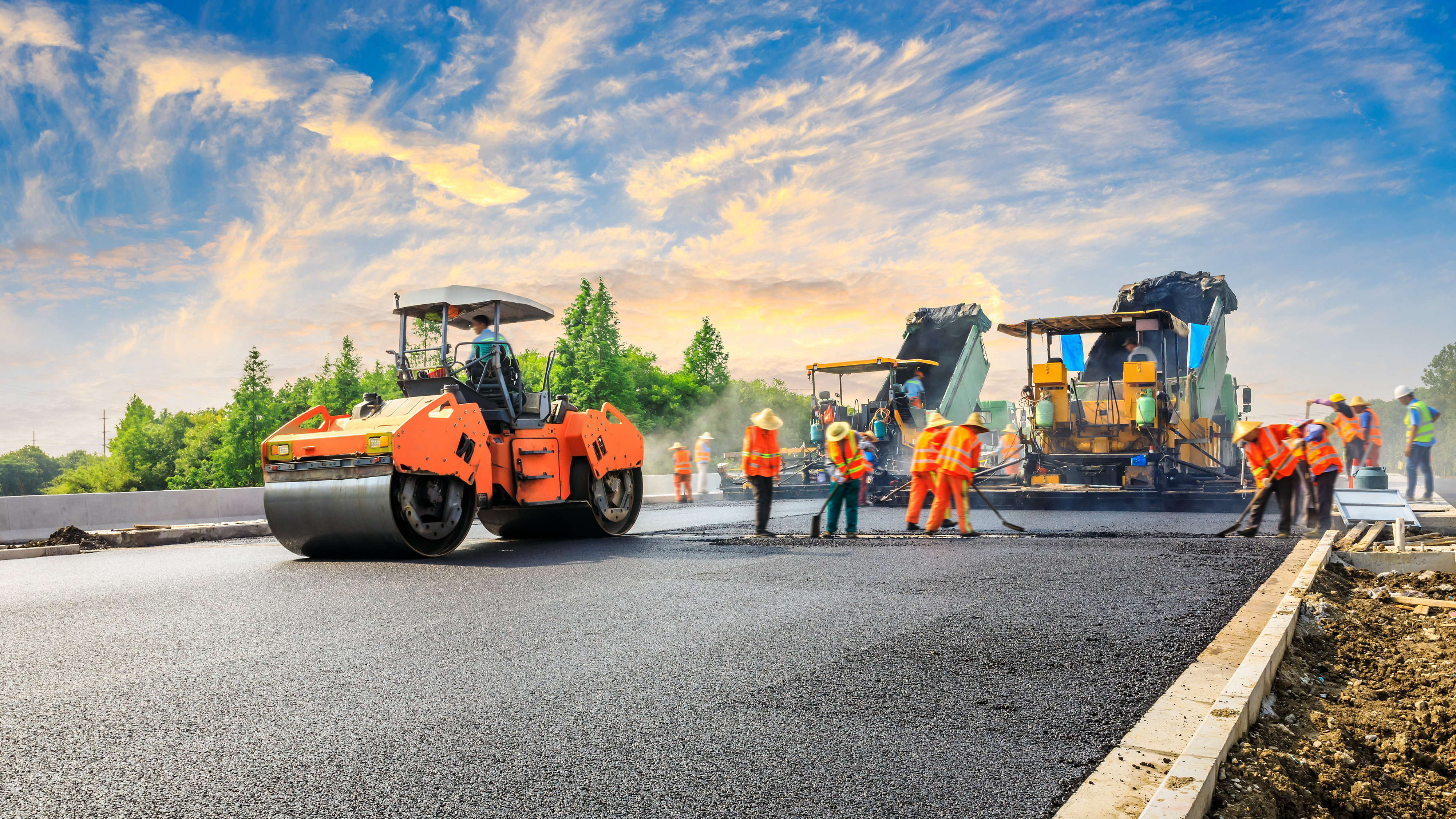 A steam roller rolling over freshly laid tarmac for a road. Workers to the right putting down more tarmac, and dump trucks in the background holding more tarmac