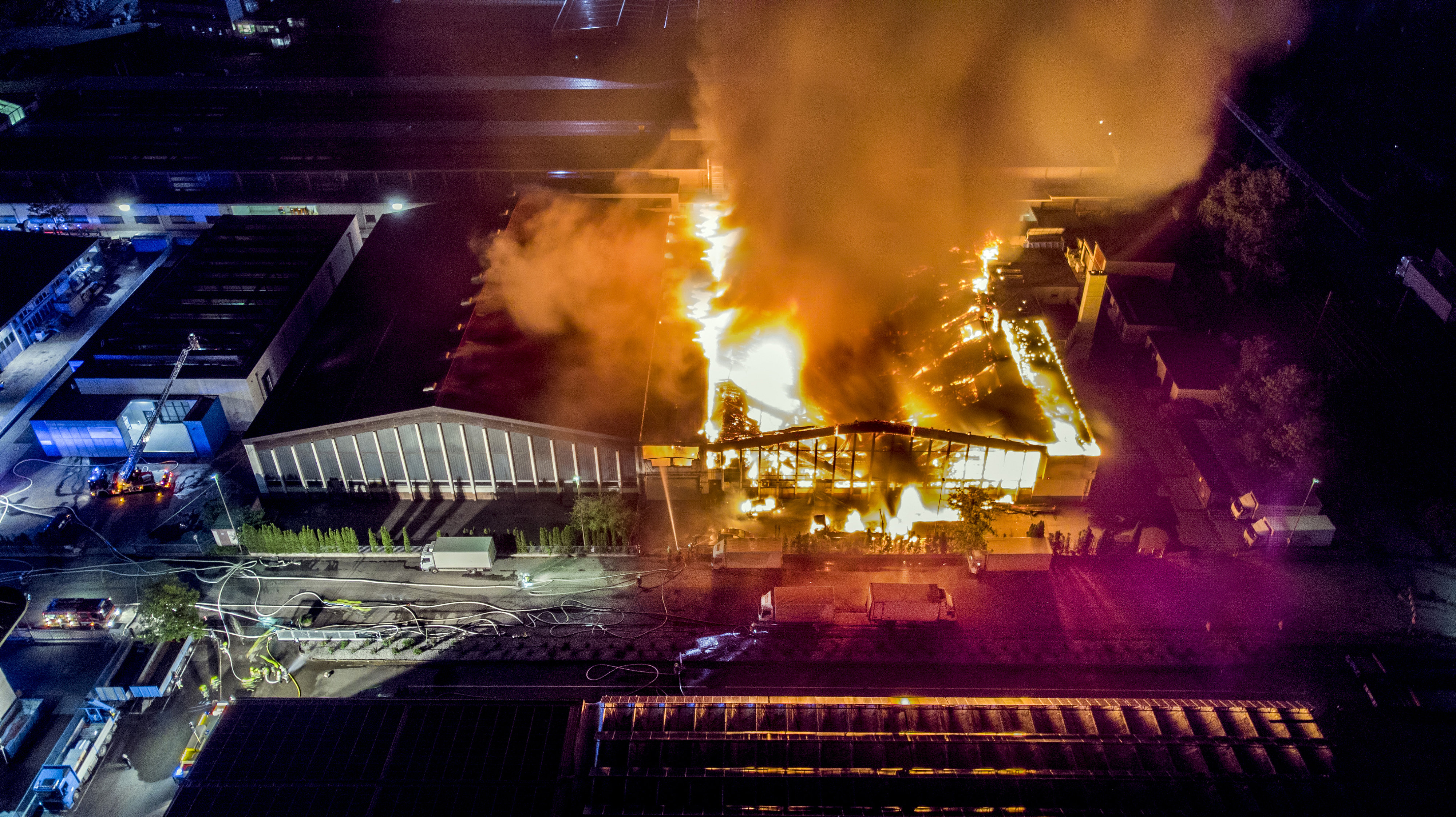 Aerial view of two warehouses, one building on fire with fire service in attendance