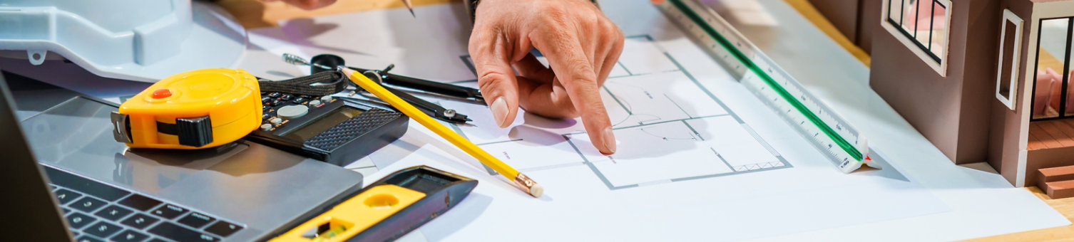 A Caucasian male engineer and an Italian male building contractor are planning and talking at a desk, examining house plans and a scale model with wooden textures for a renovation project.