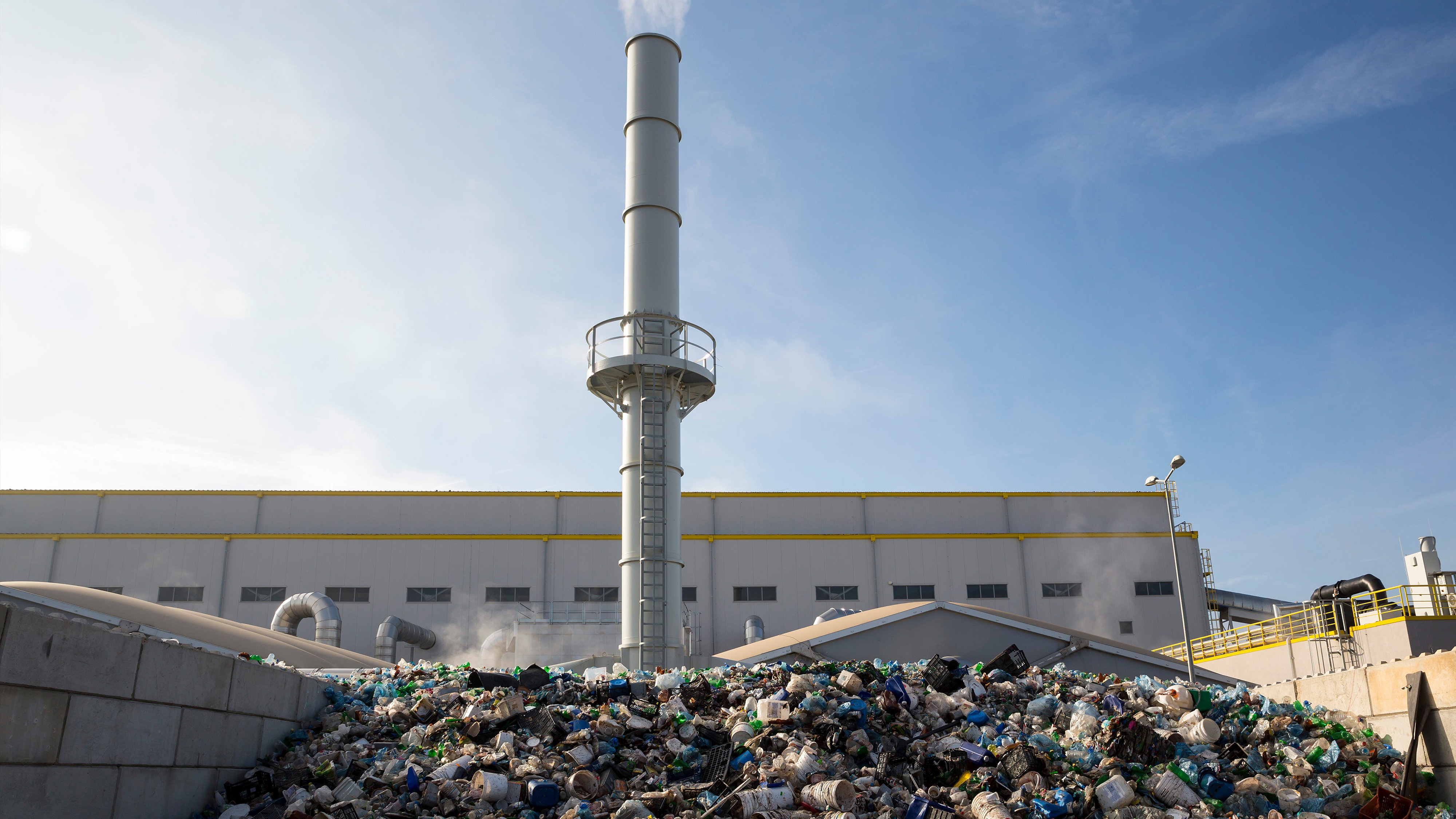 Outside of a waste to energy plant, rubbish to be burned in the foreground of the yard, and a chimney in the background.