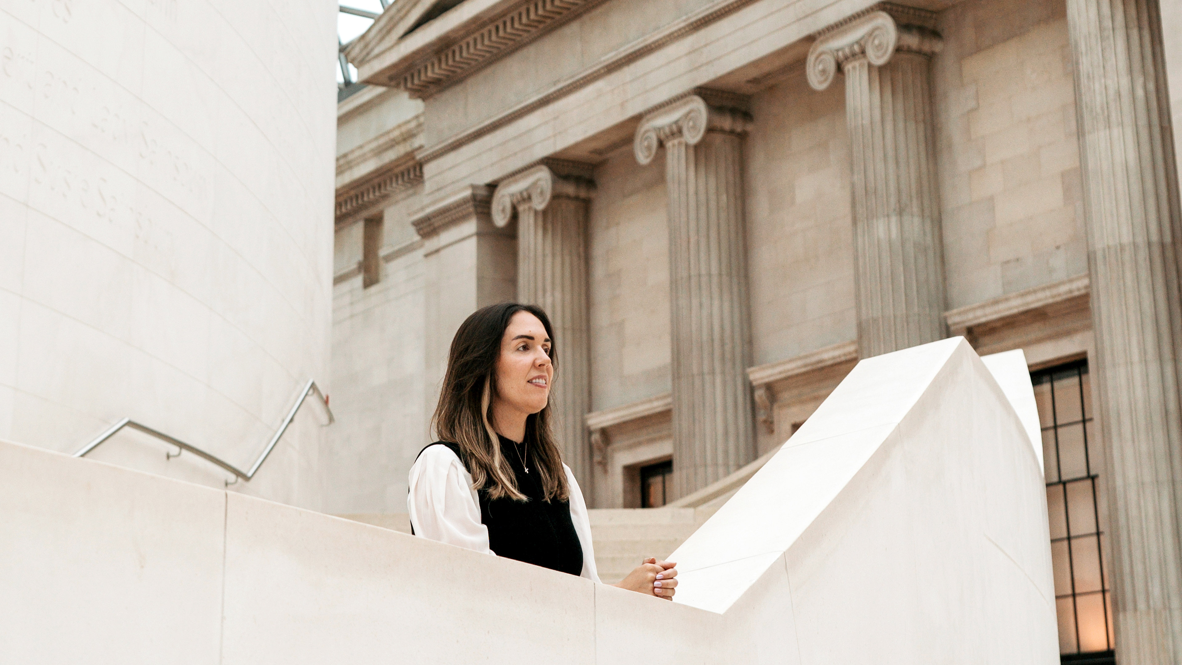 Katharine leans on ledge of marble staircase with greek columns in the background