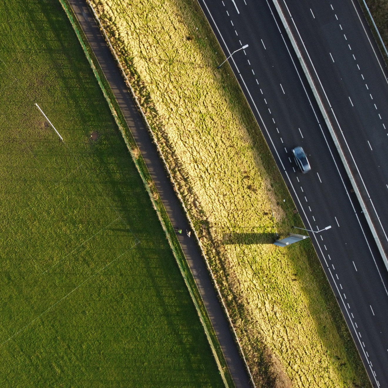 Overhead aerial photography of a public football field and a highway separated by a cycle path with cyclist and a car passing