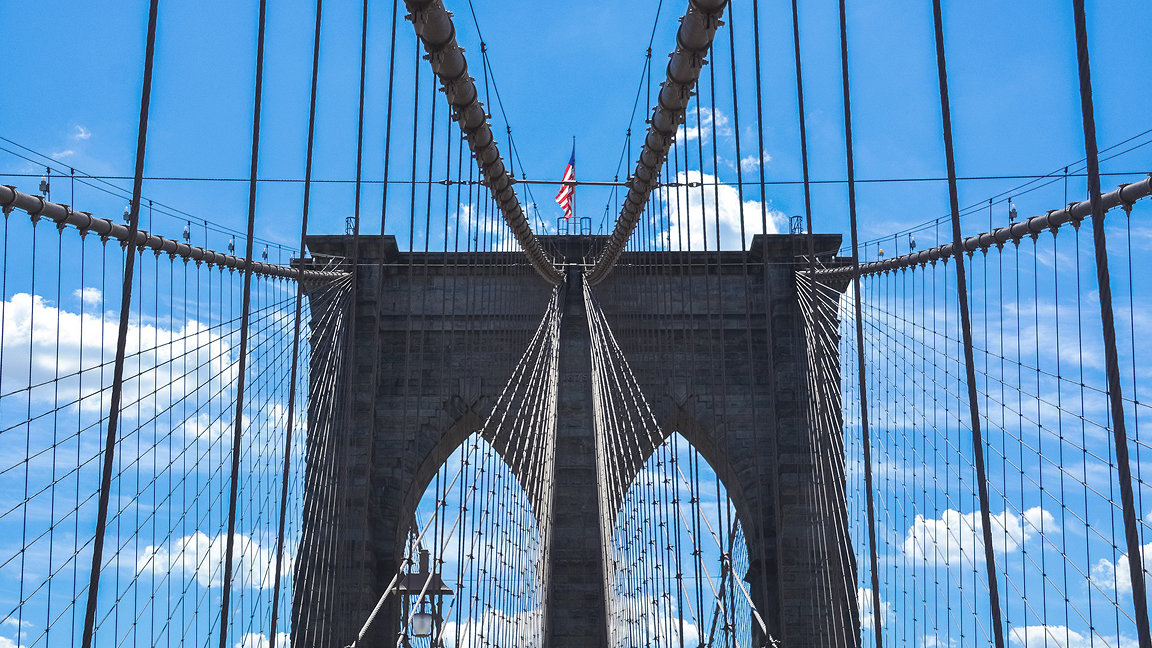 Looking front on at Brooklyn Bridge with the American flag flying and a bright blue sky in the background