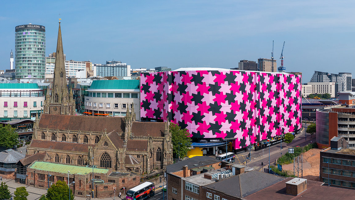 View of building covered in pink design next to a church in city skyline
