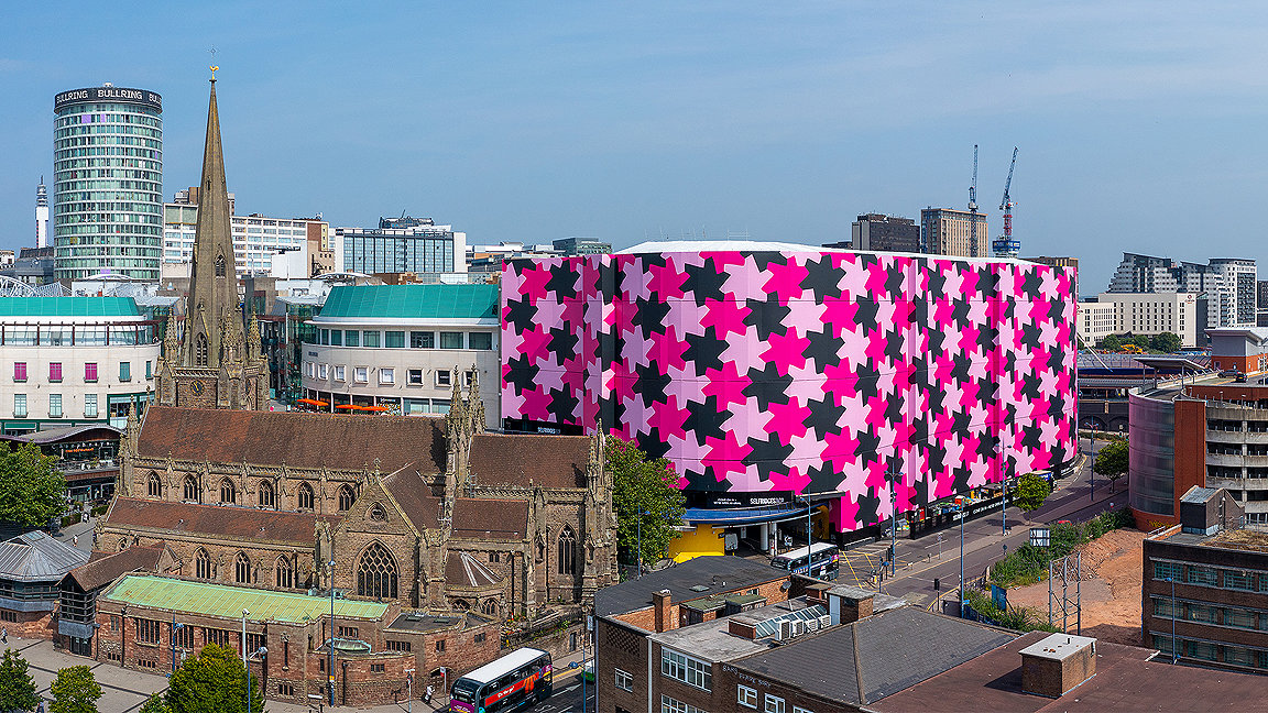 View of building covered in pink design next to a church in city skyline