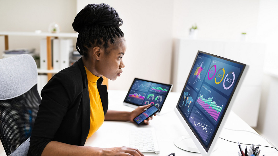 Black woman sits at desk analysing data on computer, tablet and phone