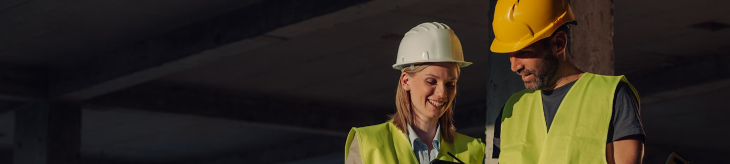 Two construction workers, a woman and a man, wearing safety vests and helmets are using a digital tablet and spirit level while working together on a building site