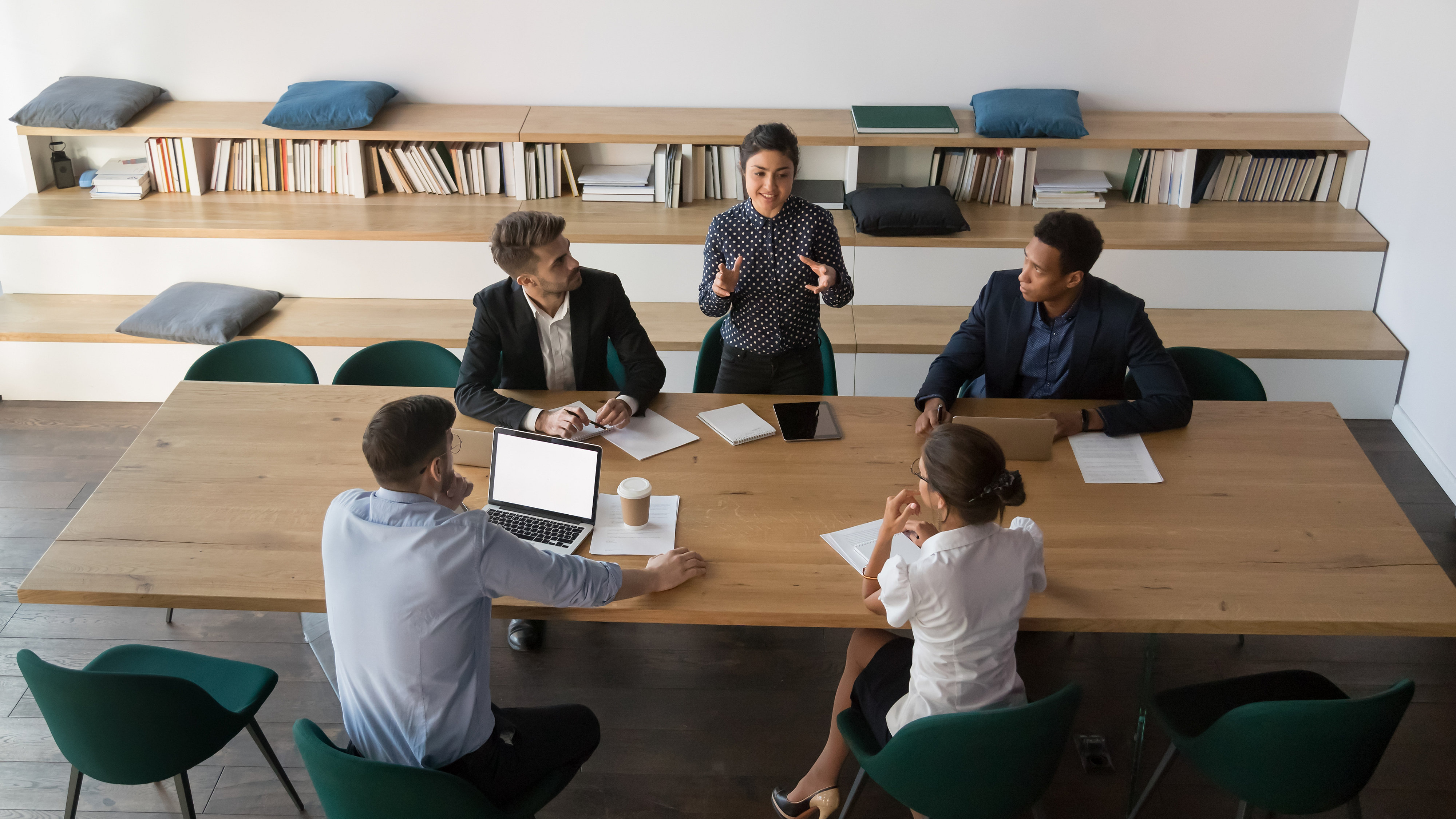 Angled shot of five professionals at a table, one is explaining something while the others watch and take notes