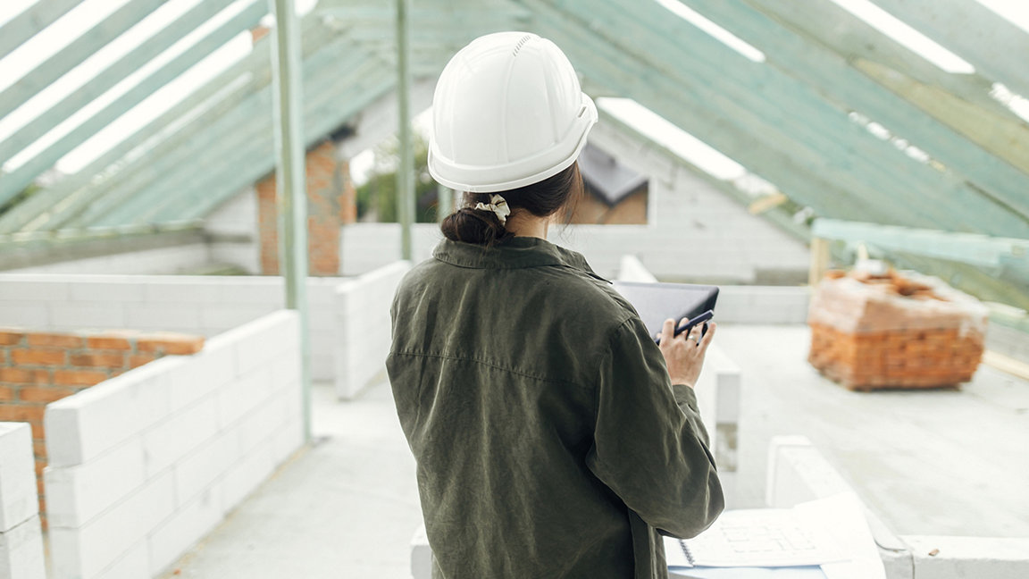 Young female architect with tablet checking blueprints against wooden roof framing of modern farmhouse.