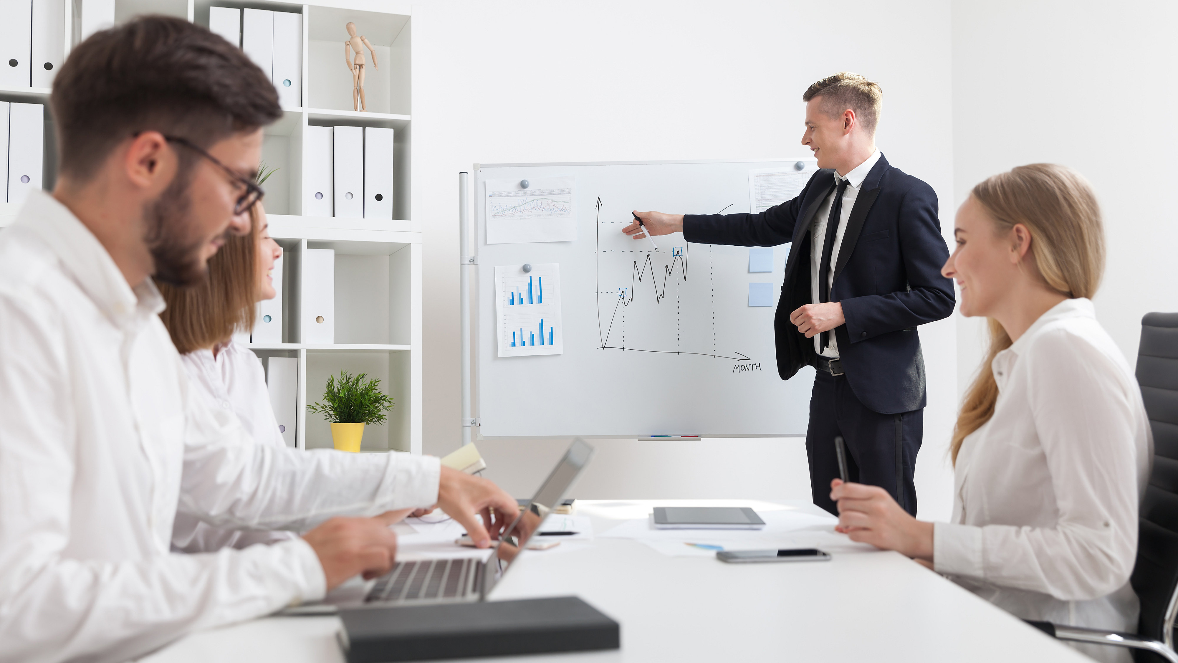 Man standing at whiteboard giving presentation in the background while male and female colleagues at desk in foreground listen