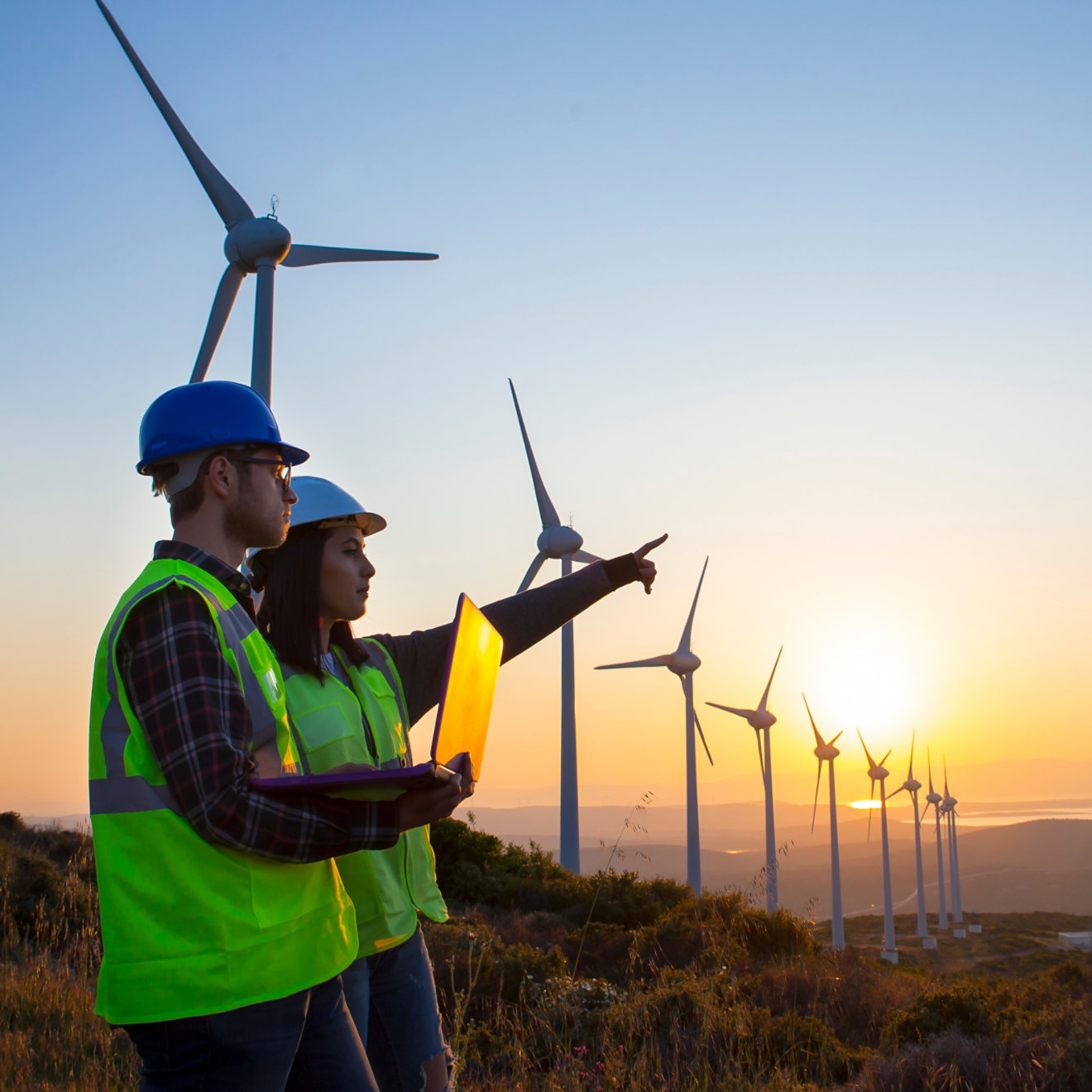 Surveyors in hi-vis vests holding a laptop outdoors at a wind farm.