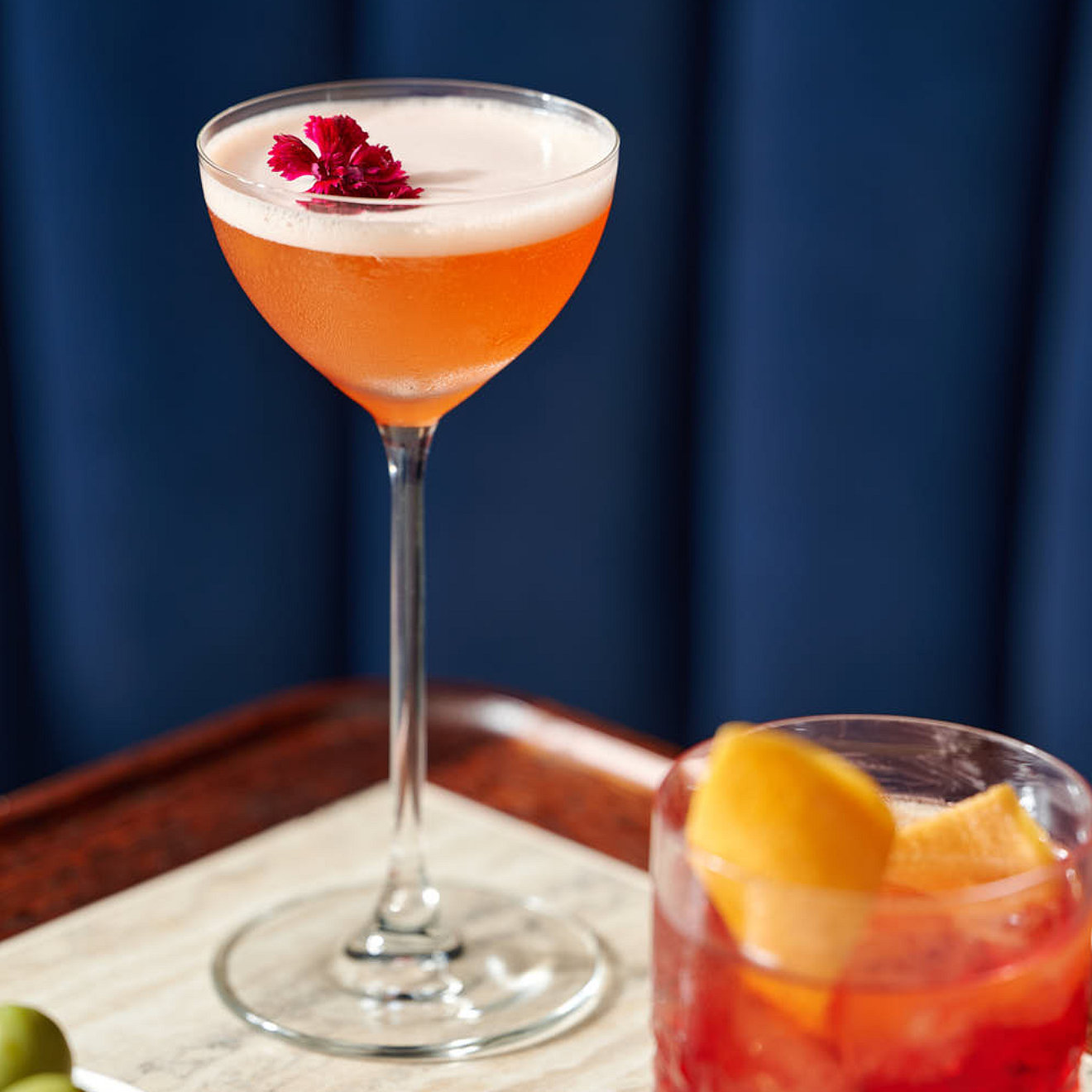 Two cocktails on a table. One is an orange cocktail with white foam. The other is red and has a twist of orange peel.