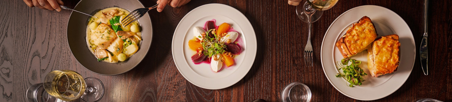 Six plates are shown from above. They have a variety of tasty looking food on them. There are four diners whose hands can be seen holding cutlery. Tehre are glasses of wine on the table.