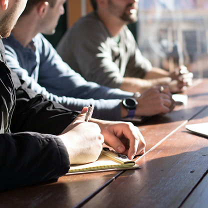 A group of men at a wooden meeting table. One is taking notes.