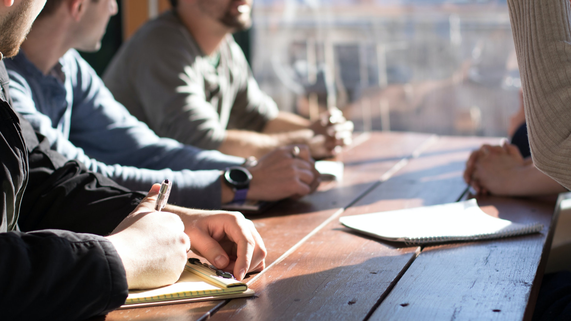 A group of men at a wooden meeting table. One is taking notes.