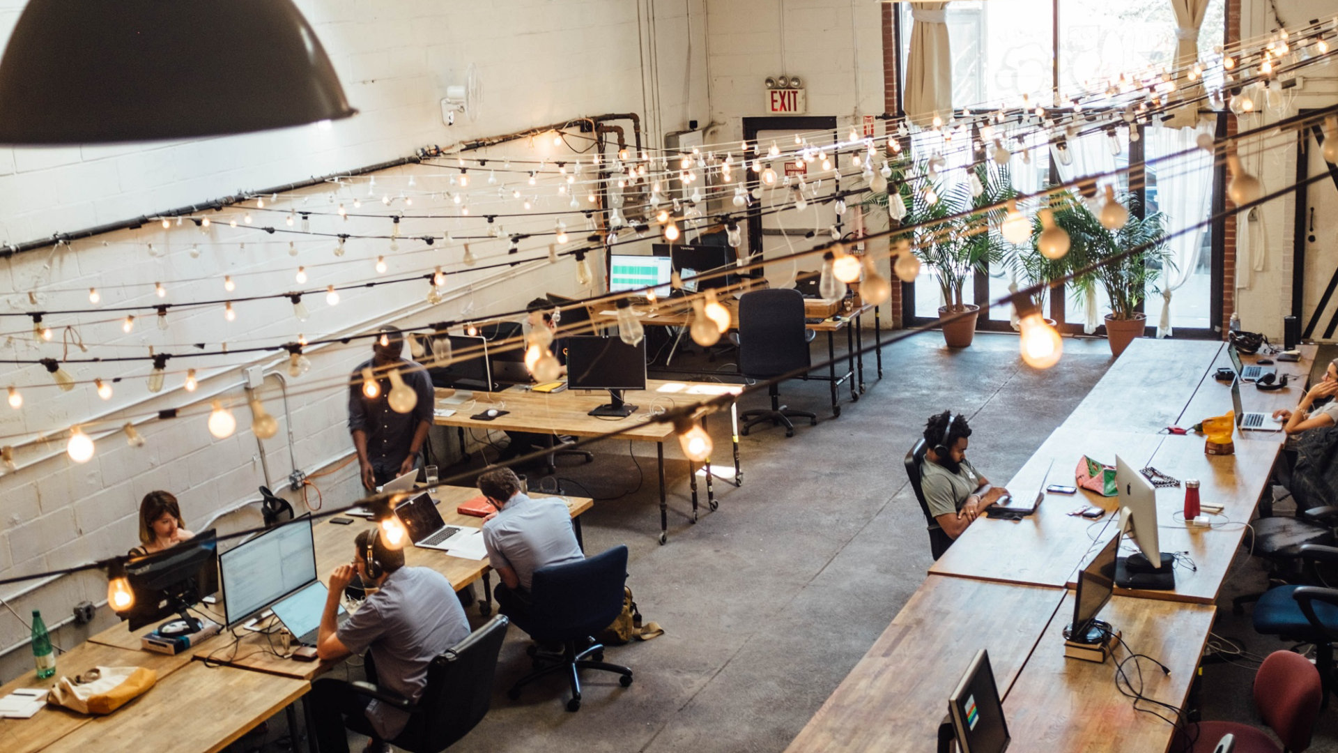 A photo of a modern office environment with wooden desks. Strings of lights run overhead.