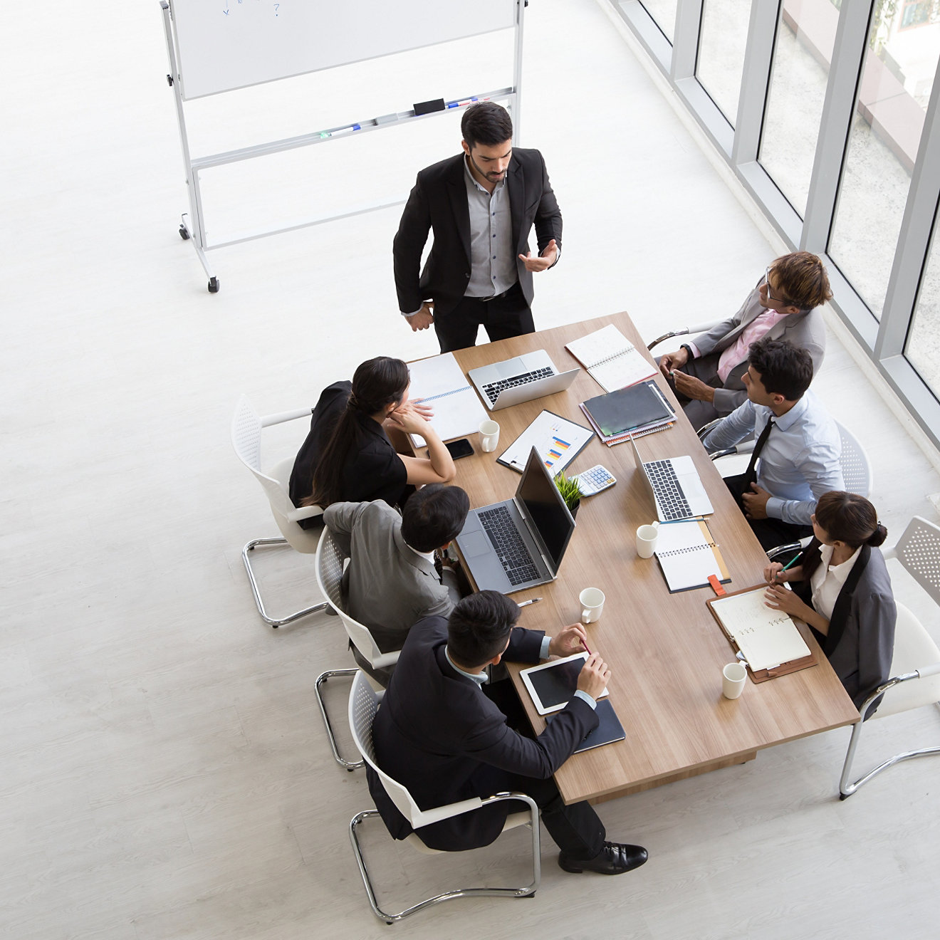 A group of young professionals in a business environment as seen from above during a meeting. One is standing and addressing the group.