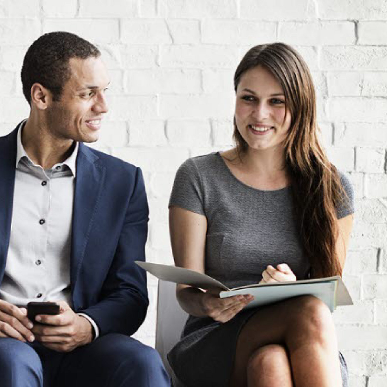 A man and woman sitting on chairs. The woman has an open folder and they are both smiling.